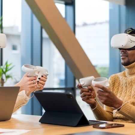 Business conference in VR in an office. A black man and caucasian woman using VR glasses and controllers, papers and gadgets on the table